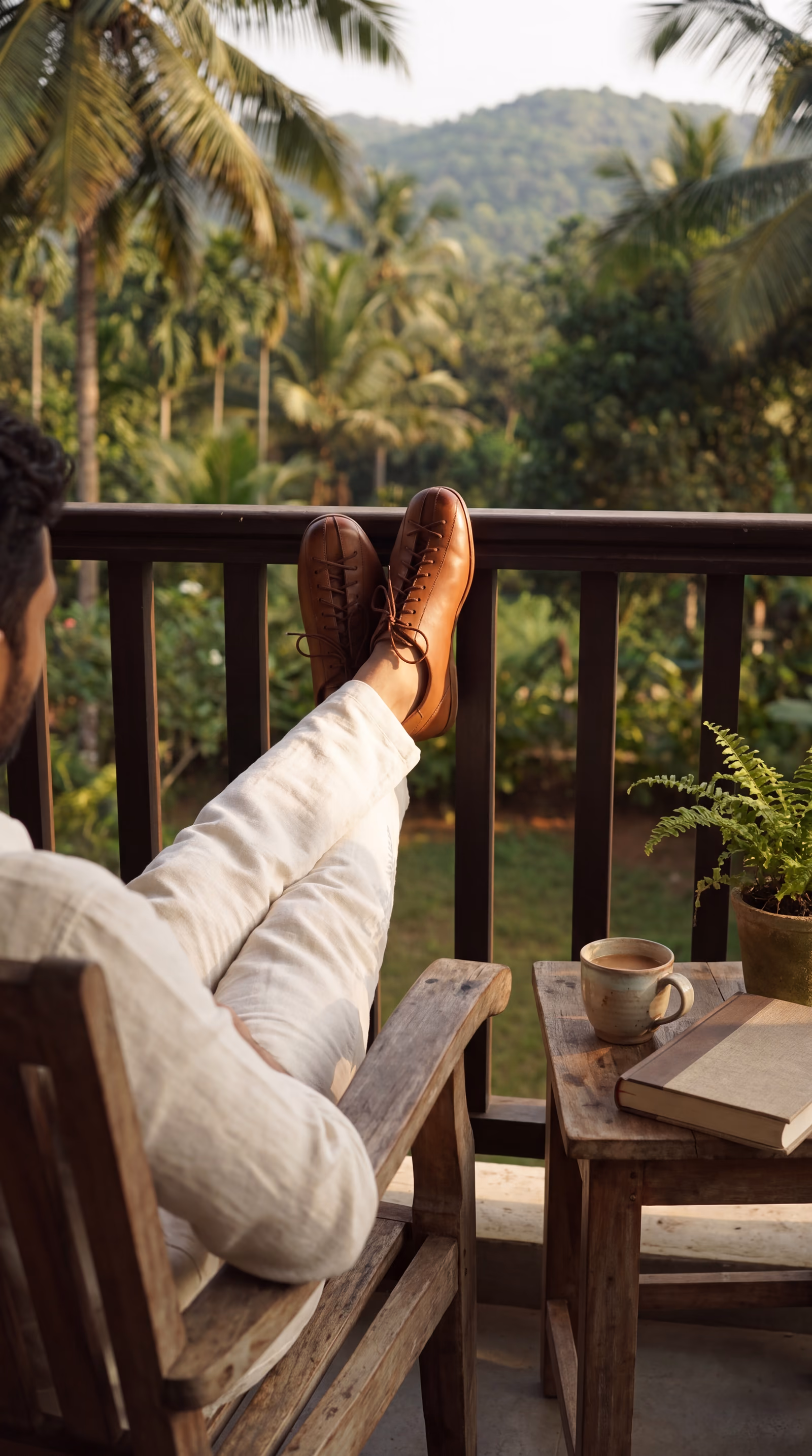 A man resting in a balcony chair with his feet up on the railing, in tan leather Kaalcari shoes, palms and hills beyond
